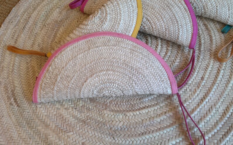 May include: Two half-circle woven straw bags with pink and yellow trim and leather handles. The bags are lying on a woven straw surface.
