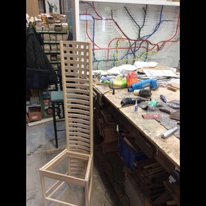 May include: A wooden chair frame in a workshop setting. The chair is made of light wood and has a slatted back and seat. The chair is unfinished and is sitting on a workbench.