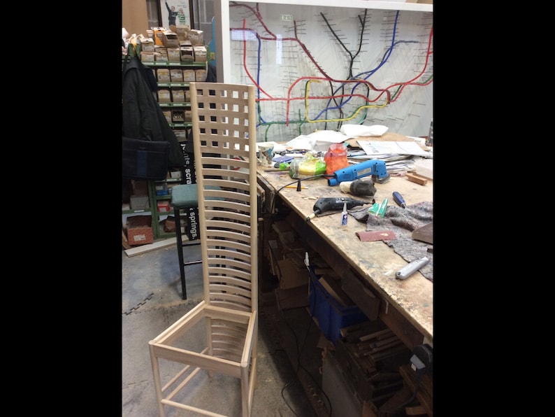 May include: A wooden chair frame in a workshop setting. The chair is made of light wood and has a slatted back and seat. The chair is unfinished and is sitting on a workbench.