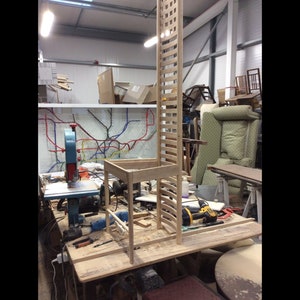 May include: A wooden chair with a tall back is being assembled in a workshop. The chair is made of light-colored wood and has a simple design. The chair is sitting on a workbench with tools and other woodworking materials around it.