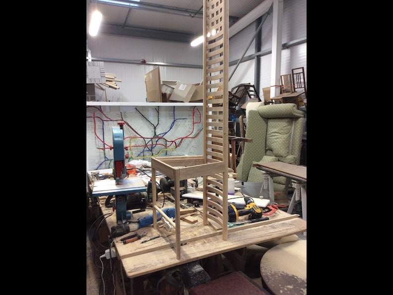 May include: A wooden chair with a tall back is being assembled in a workshop. The chair is made of light-colored wood and has a simple design. The chair is sitting on a workbench with tools and other woodworking materials around it.