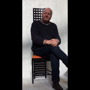 May include: A man sits in a black and orange chair with a geometric design. The chair has a high back and a wide seat. The man is wearing a dark blue sweater and jeans.