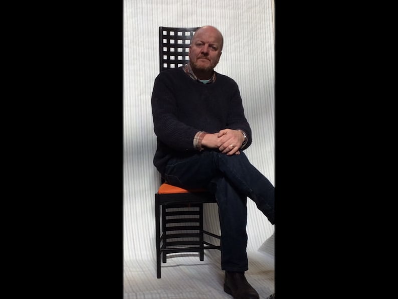 May include: A man sits in a black and orange chair with a geometric design. The chair has a high back and a wide seat. The man is wearing a dark blue sweater and jeans.