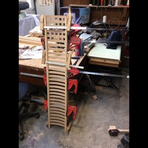 May include: Two wooden chair backs are being clamped together to form a curved shape. The chair backs are made of thin strips of wood and are being held together with red clamps. The chair backs are sitting on a wooden table in a workshop.