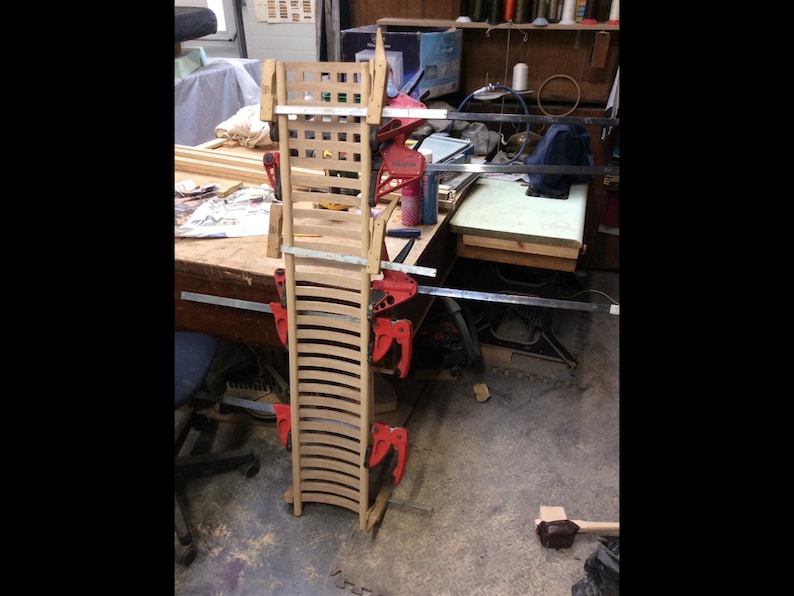 May include: Two wooden chair backs are being clamped together to form a curved shape. The chair backs are made of thin strips of wood and are being held together with red clamps. The chair backs are sitting on a wooden table in a workshop.