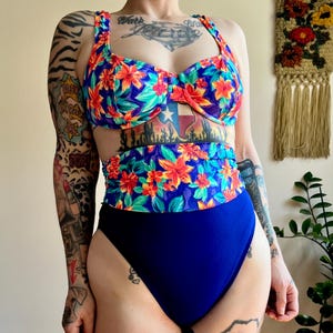May include: A two-piece swimsuit with a vibrant floral pattern. The top features a bustier design with a sweetheart neckline, and the high-waisted bottoms are a solid navy blue. The floral print includes red, orange, and green flowers on a blue background.