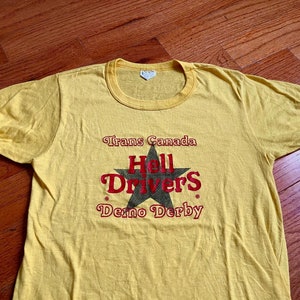 May include: Yellow t-shirt with a black star and the text "Trans Canada Hell Drivers Demo Derby".