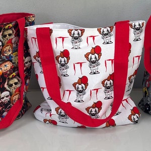 May include: Three tote bags featuring different horror movie character designs. The first bag has a black background with colourful horror movie characters. The second bag has a white background with a repeating pattern of a clown with a red balloon and the word "IT". The third bag has a grey background with a variety of horror movie characters.