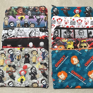 May include: A collection of rectangular fabric zip pouches featuring various horror movie character prints. The designs include chibi-style illustrations of iconic villains, with one pouch specifically displaying the text Chucky He wants you as a best friend.
