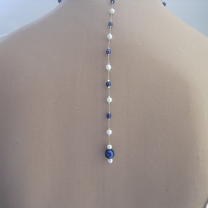 Può includere: Una lunga collana delicata con perle bianche e blu scuro alternate. La collana è infilata su una sottile catena d'argento e pende sul retro del collo di una persona.