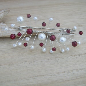 Peut inclure: Une broche en fil d'argent avec des perles blanches et bordeaux. La broche a la forme d'une explosion de lumière.