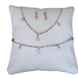 Könnte beinhalten: Ein Schmuckset mit Halskette, Armband und Ohrringen. Das Set umfasst eine silberfarbene Kette mit rosa Perlen und einem Schmetterlingsanhänger. Die Ohrringe sind rosa Perlenstecker. Der Schmuck wird auf einem weißen Kissen präsentiert.