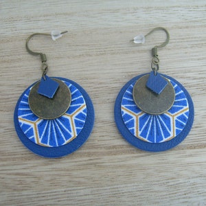 Könnte beinhalten: Ein Paar handgefertigte Ohrringe mit einem mehrschichtigen Design. Jeder Ohrring hat eine blaue Lederbasis, eine Stoffschicht mit blauem und goldenem geometrischem Muster und eine bronzefarbene Metallscheibe. Obenauf befindet sich ein kleines blaues Lederdiamant.