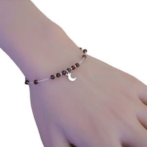 Puede incluir: Una pulsera delicada con pequeñas cuentas de color rojo oscuro ensartadas en un cordón fino de color claro. Un dije de media luna plateada cuelga del centro. La pulsera se muestra en la muñeca.