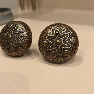 Könnte beinhalten: Zwei verzierte Messing-Schrankknöpfe mit sternförmigem Design und komplizierten floralen Mustern.