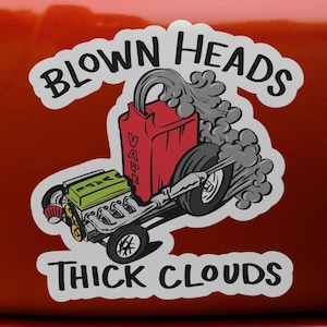 Puede incluir: Una pegatina que muestra un motor de dibujos animados con un contenedor de combustible rojo etiquetado como VAPE, emitiendo nubes de humo gris. El texto BLOWN HEADS está arqueado arriba y THICK CLOUDS impreso abajo.