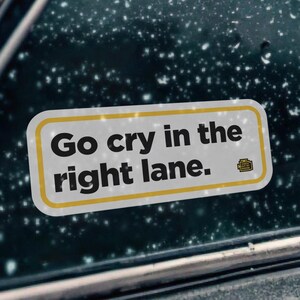 Puede incluir: Una pegatina rectangular para coche con fondo blanco y borde amarillo. El adhesivo presenta el texto negro "Go cry in the right lane." seguido de un pequeño icono de motor amarillo. Está pegado en una ventanilla oscura cubierta de gotas de lluvia.