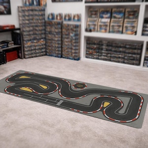 May include: A rectangular gray play mat featuring a race track design. The track is black with yellow and white markings, and red and white striped borders. The mat is designed for toy cars and racing games.
