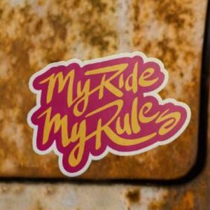 以下が含まれることがあります： ピンクと黄色の「My Ride My Rules」という文字が入ったステッカーが貼られた、ヴィンテージカーの後部セクションのクローズアップ。車のボディには錆が見られ、テールライトも確認できます。