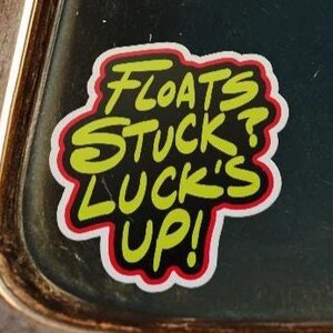 Puede incluir: Una pegatina con el texto "FLOATS STUCK? LUCK'S UP!" en amarillo, delineado en negro y rojo. La pegatina está sobre una superficie oscura, posiblemente una ventana de coche.