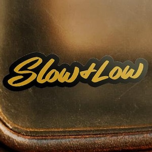 Puede incluir: Una pegatina negra y dorada con las palabras "Slow + Low" en una fuente cursiva. La pegatina está sobre una superficie marrón texturizada, posiblemente un vehículo. El texto es dorado con un contorno negro.