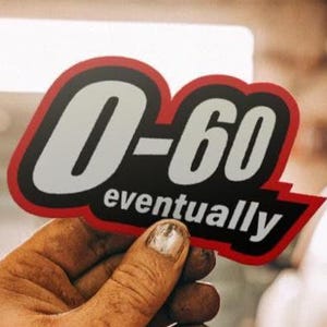 May include: A sticker with the text "0-60 eventually" in a black, red, and white design. The sticker is held up by a hand with dirty fingers. The background is blurred, suggesting a garage or workshop setting.
