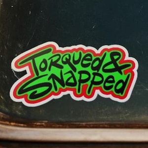 Puede incluir: Una pegatina con las palabras "Torqued & Snapped" en verde, delineadas en rojo y blanco. La pegatina está sobre una superficie oscura, posiblemente la ventana de un coche.