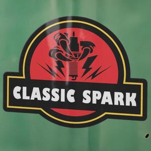 May include: A circular sticker with a red background and black border applied to a sage green surface. The sticker features a black graphic of a spark plug with lightning bolts and the white text CLASSIC SPARK centred in a black rectangular banner.