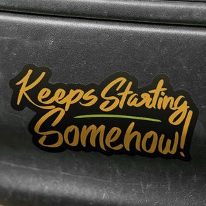 Puede incluir: Una pegatina negra con el texto "Keeps Starting Somehow!" en letras doradas y verdes. La pegatina está adherida a un parachoques de coche negro, mostrando un mensaje humorístico.
