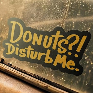 Puede incluir: Una calcomanía de vinilo troquelada aplicada sobre una superficie de vidrio, con el texto amarillo "Donuts?! DisturbMe." en una fuente negrita y estilizada con un borde negro. El adhesivo está colocado en una ventana, mostrando un mensaje claro.