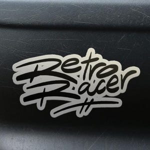 以下が含まれることがあります： スタイリッシュなフォントで「Retro Racer」という言葉が書かれた白いステッカーが、濃い灰色の車のバンパーに貼られています。ステッカーは黒いアウトラインとハッシュタグ記号が付いています。