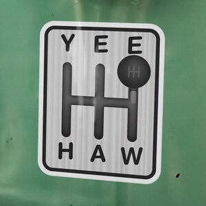 Puede incluir: Una pegatina rectangular con un gráfico en blanco y negro de un patrón de cambio de marchas manual. El texto YEE está impreso sobre el patrón y HAW debajo. Un pequeño icono negro de pomo de palanca de cambios se sitúa en la marcha superior derecha.
