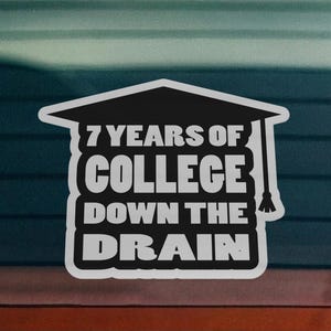 May include: A black and white vinyl decal featuring the shape of a graduation cap with a tassel. The text printed in white reads 7 YEARS OF COLLEGE DOWN THE DRAIN, arranged in four lines within the silhouette of the cap on a car window.