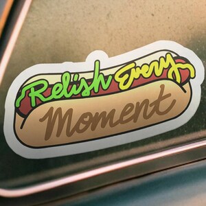 以下が含まれることがあります： 漫画のホットドッグと「Relish Every Moment」の文字が入ったステッカー。 ホットドッグにはパン、レリッシュ、マスタードが入っています。 ステッカーは白で、わずかな影があります。