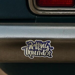 Puede incluir: Una pegatina con las palabras "Rolling Thunder" en una fuente estilizada. El texto es azul con un contorno blanco, sobre un fondo plateado. La pegatina está adherida a un vehículo.