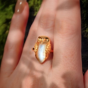Könnte beinhalten: Ein goldfarbener Eulenring mit einem perlmuttfarbenen weißen Mittelstein. Der Ring weist detaillierte Federstrukturen auf und ist auf einem dünnen Goldband gefasst. Die Augen und der Schnabel der Eule sind deutlich zu erkennen.