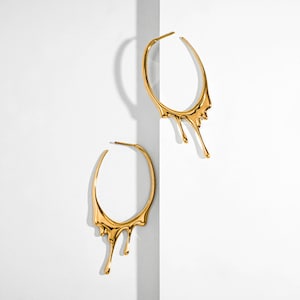 May include: A pair of gold hoop earrings with a dripping design. The earrings are made of a smooth, shiny metal and have a modern, abstract look.