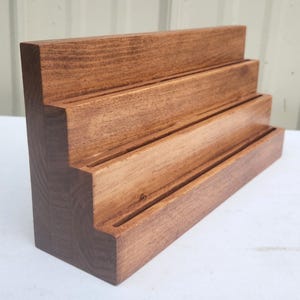 May include: A tiered wooden display stand featuring three distinct levels with horizontal grooves carved into each step. The stand is crafted from natural brown wood with a visible grain pattern, showcasing a smooth finish and a stepped design.