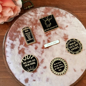 May include: A collection of six enamel pins with black and gold accents. The pins feature various quotes and designs, including a clock face, a snake, and a hand holding another hand. The pins are displayed on a pink velvet background.