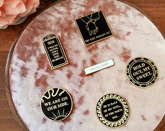May include: A collection of six enamel pins with black and gold accents. The pins feature various quotes and designs, including a clock face, a snake, and a hand holding another hand. The pins are displayed on a pink velvet background.