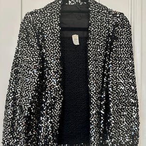 May include: A black sequin jacket hanging on a black hanger. The jacket is covered in small, shiny sequins, creating a textured, shimmering effect. The interior lining is black, and the jacket has a classic open-front design.
