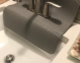 Könnte beinhalten: Eine graue, rechteckige und texturierte Silikon-Spritzschutzmatte für Wasserhähne, die um die Basis einer gebürsteten Nickel-Armatur im Badezimmer liegt. Die Matte hat einen mittigen Ausschnitt für die Griffe und den Auslauf des Wasserhahns.
