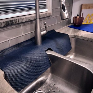 May include: A navy blue sink splash guard made of a textured material, designed to fit around a kitchen faucet. The guard sits on the countertop next to a stainless steel sink, protecting the area from water splashes.