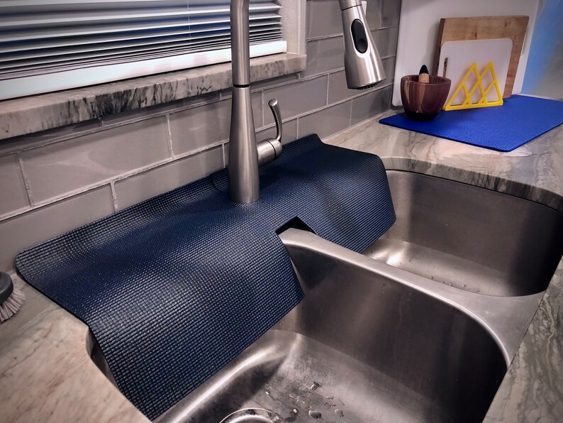 May include: A navy blue sink splash guard made of a textured material, designed to fit around a kitchen faucet. The guard sits on the countertop next to a stainless steel sink, protecting the area from water splashes.