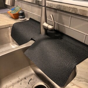 May include: A black sink mat with a textured surface is draped over a stainless steel sink. The mat is designed to protect the countertop from water damage. A silver faucet is centered above the mat.