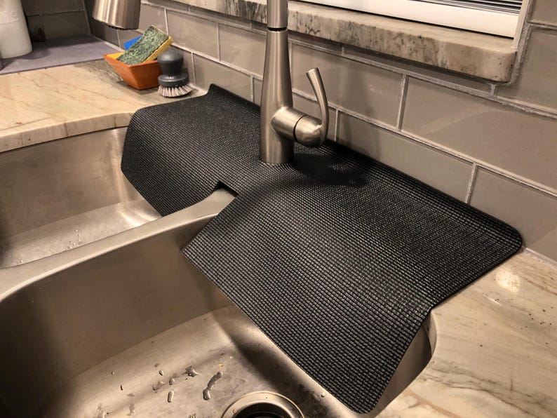 May include: A black sink mat with a textured surface is draped over a stainless steel sink. The mat is designed to protect the countertop from water damage. A silver faucet is centered above the mat.