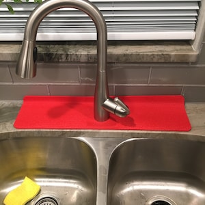May include: A stainless steel kitchen faucet with a high arc spout is positioned above a double sink. A bright red mat sits behind the faucet. A yellow sponge rests in the left sink basin.