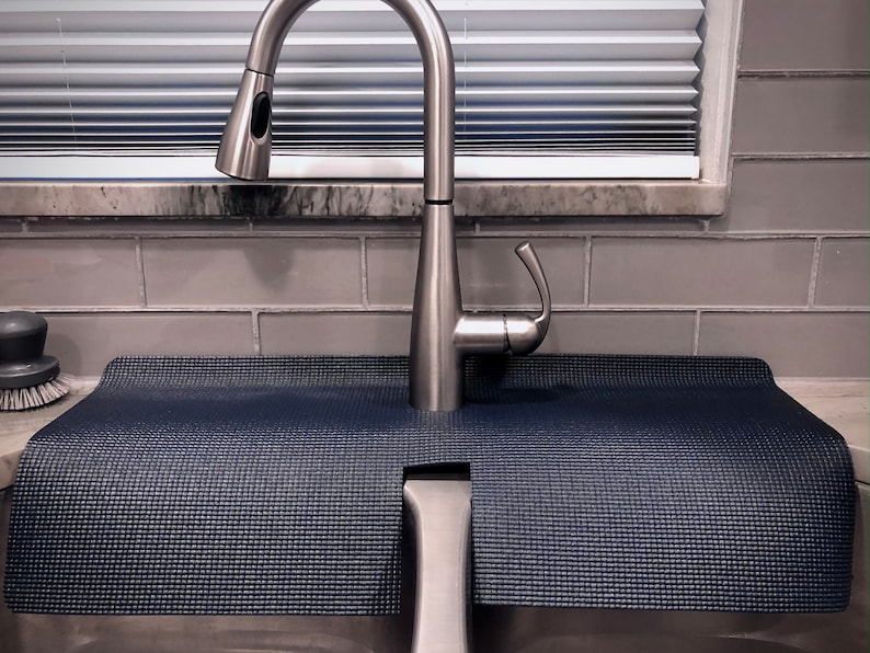 May include: A navy blue sink mat with a textured surface, designed to fit around a kitchen faucet. The mat is positioned in front of a stainless steel faucet, with a gray tile backsplash and a window with blinds in the background.