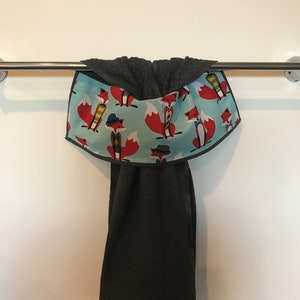 May include: A dark gray towel with a matching hood, hanging from a metal bar. The hood features a light blue fabric printed with red foxes wearing hats and scarves. The towel is made of a textured material.