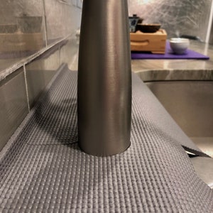 May include: A gray yoga mat with a textured surface is positioned near a stainless steel faucet. The mat is rolled out on a countertop, with a sink visible in the background. The faucet is cylindrical and has a brushed metal finish.